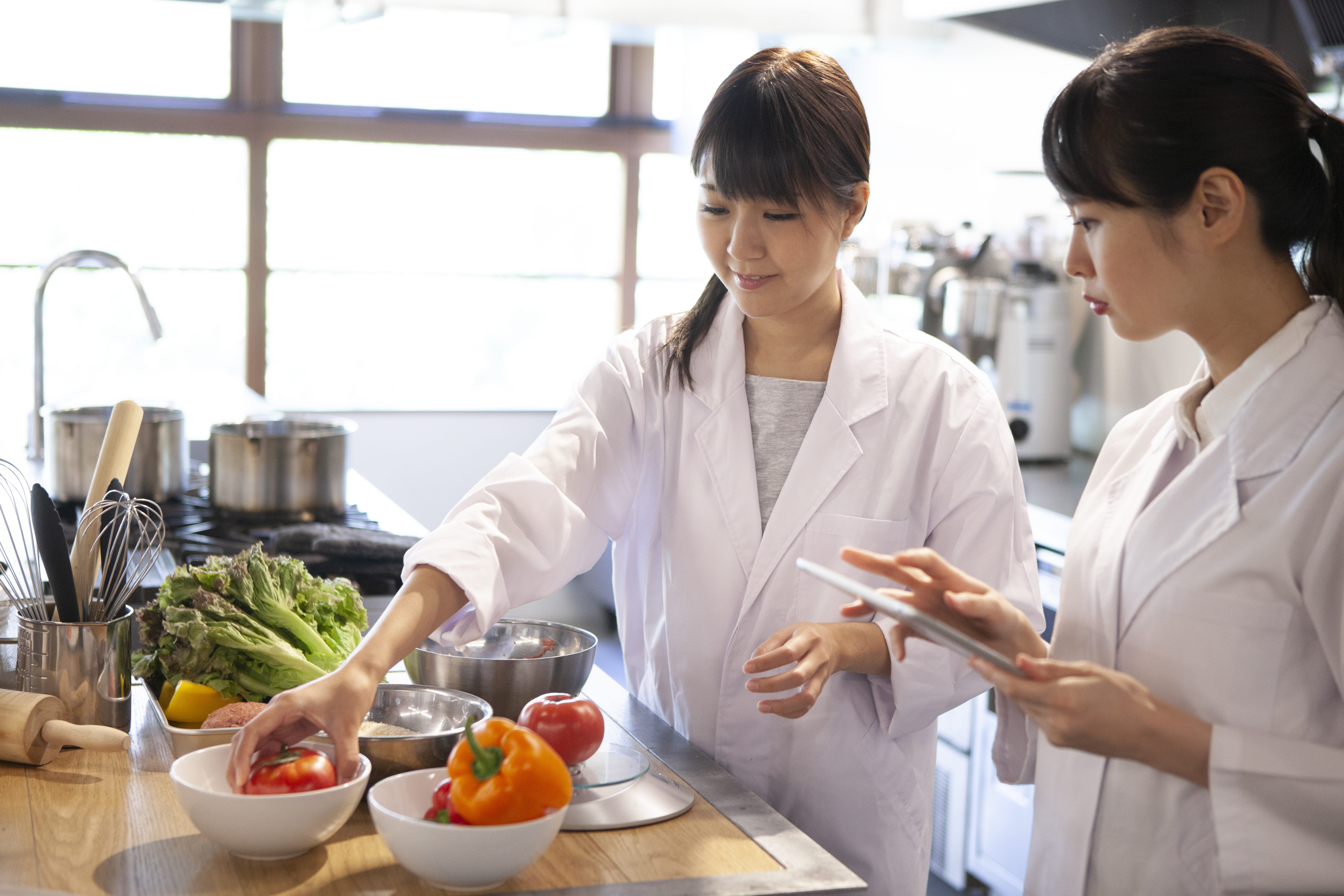 Two people in lab coats examining vegetables in bowls in an industrial kitchen.